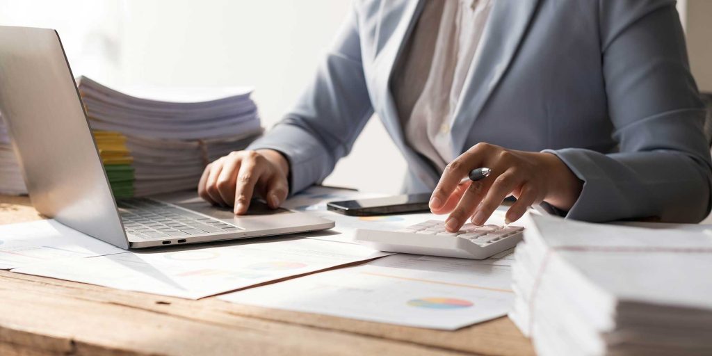 Person in a suit working at a desk with a laptop, documents, calculator, and smartphone, performing calculations and data analysis.