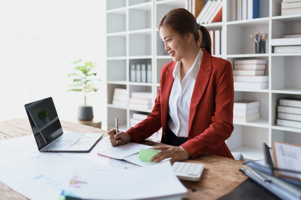 Young woman in red blazer working and writing notes at a desk with a laptop and office supplies in a bright workspace.